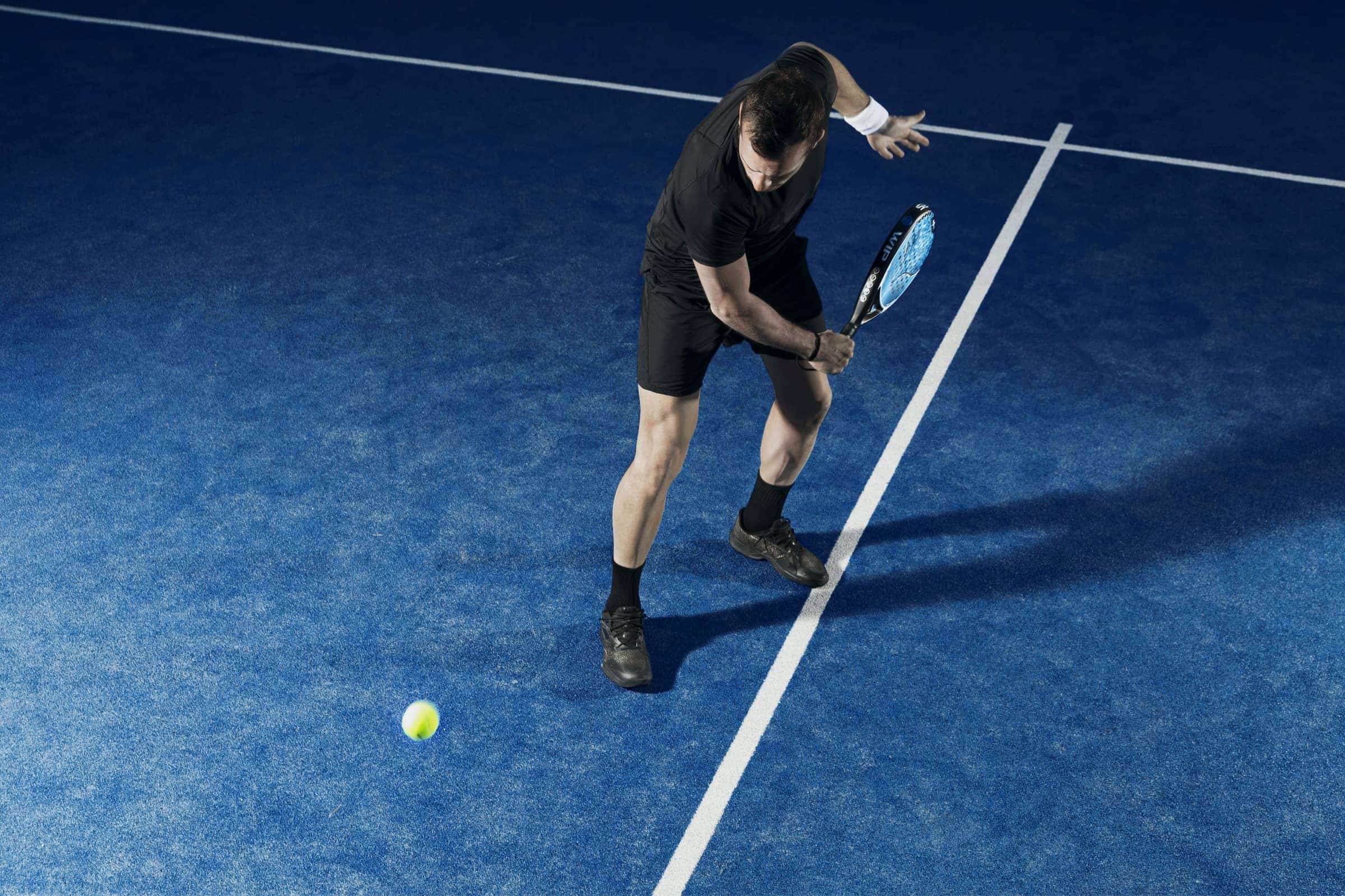 لاعب بادل بملابس سوداء ينحني ليردّ الكرة على ملعب أزرق تحت إضاءة خافتة.